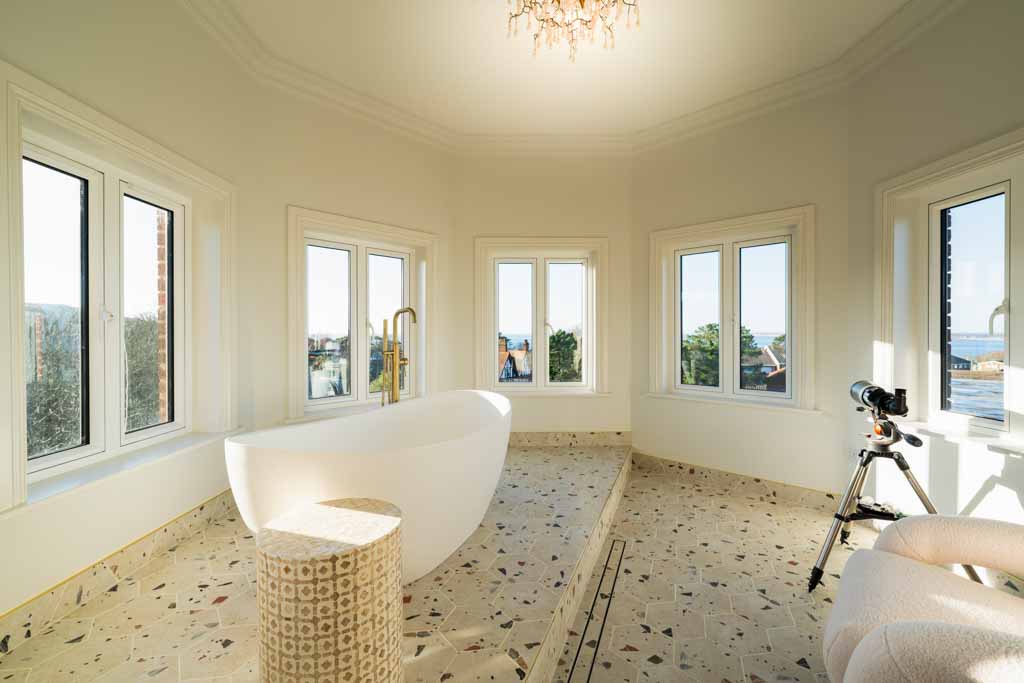 Luxurious bathroom featuring a modern freestanding white bath with a gold tap, surrounded by large windows that let in abundant natural light. The floor is covered in patterned tiles, and a small round stool sits beside the bath. A telescope on a tripod is positioned near a plush chair, suggesting a relaxing and contemplative atmosphere. The room feels serene, spacious, and inviting, with views of trees and rooftops outside.